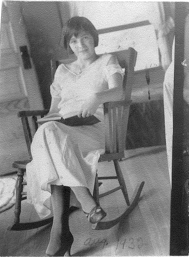 black and white picture of a white woman with dark hair in a white dress sitting in a rocking chair with a book; aug. 1932 is written in the bottom-right corner