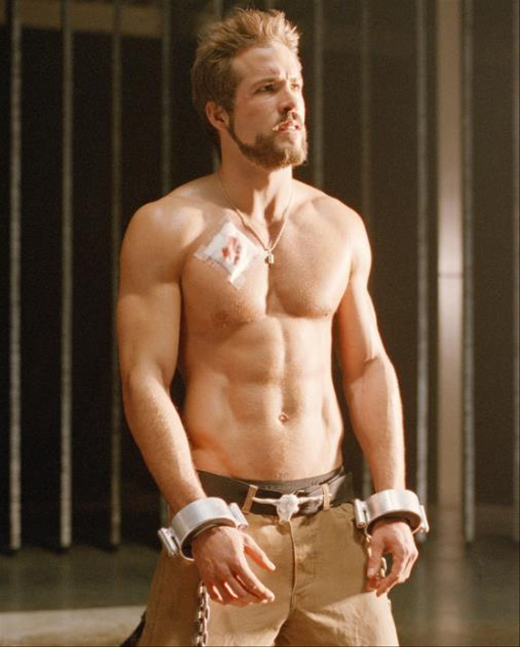 ryan reynolds, a muscular white man in khaki pants and no shirt kneels in a room with chains on his wrists and a wound on his upper shoulder