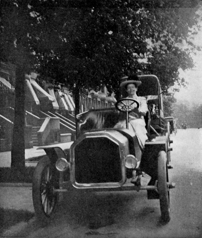 black and white picture of a white woman in a white dress and a hat driving an early nineteen-teens car