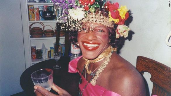 Marsha P. Johnson, a black transwoman, with bright flowers on her head, bright pink top, and  necklaces sits at a table.